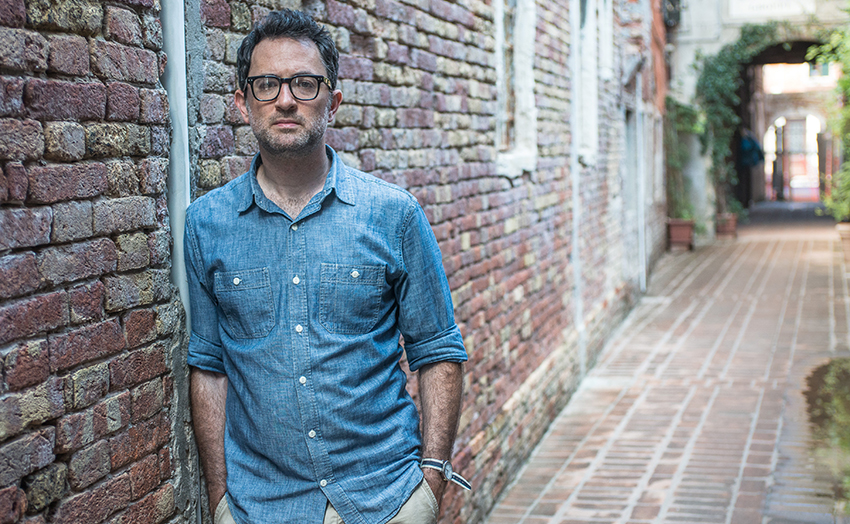 Chinan Tigay, wearing a denim work shirt with the sleeves rolled up, is standing in a cobblestone alley, leaning against a brick wall in this photo. Photo credit: Emanuele Dello Strologo